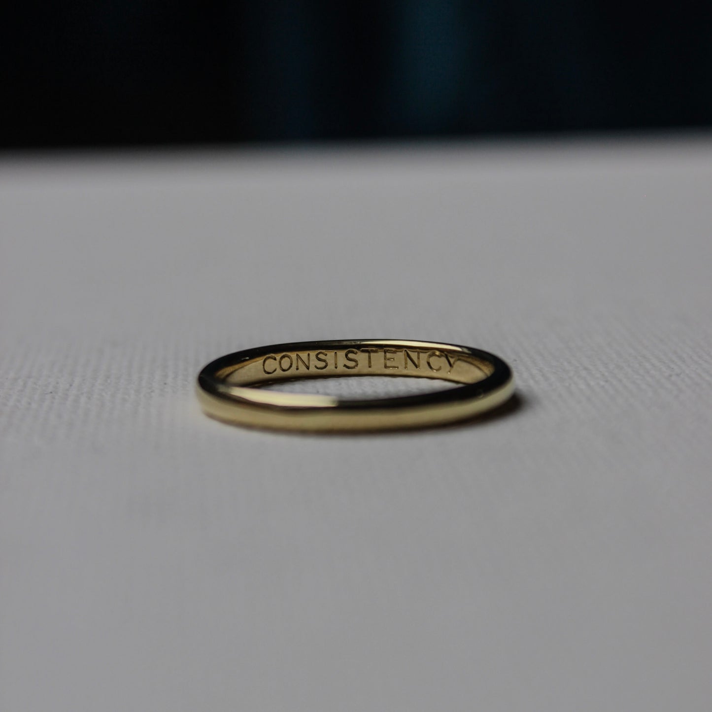 Gold ring with 'CONSISTENCY' engraving on a light gray background