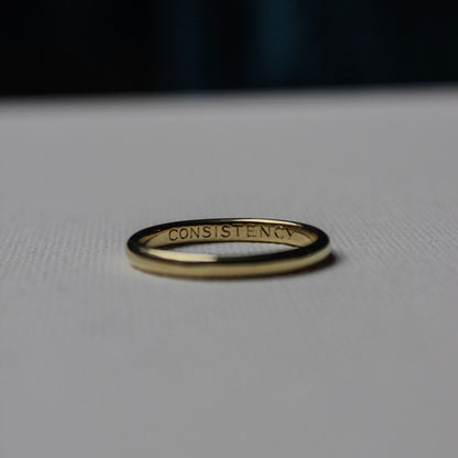 Gold ring with 'CONSISTENCY' engraving on a light gray background